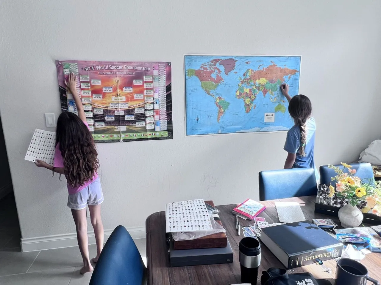 Two kids at a dining room wall, one placing a sticker on a World Cup bracket poster, the other finding a country on a world map
